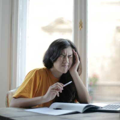 Ethnic female student in casual wear sitting at table with laptop and notebook and grimacing from migraine while doing homework during distance learning