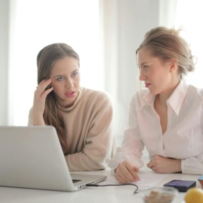 Pensive female entrepreneurs wearing casual clothes sitting together at table with laptop and talking about problems in project