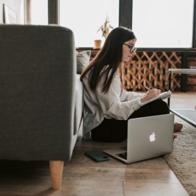 Woman Writing On Notebook With Laptop By Her Side