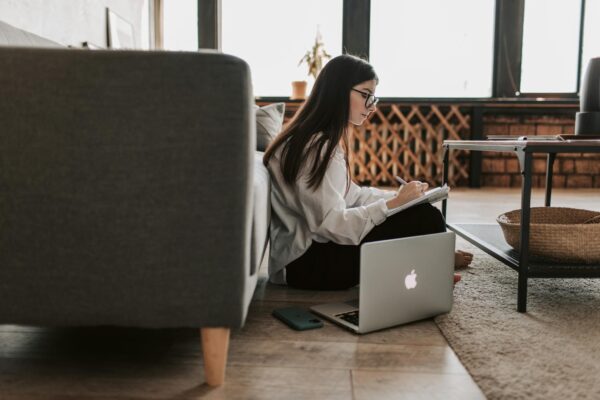 Woman Writing On Notebook With Laptop By Her Side